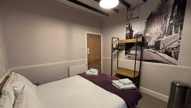 A bedroom with a bunk bed, a radiator, a door, a wall-mounted shelf with clothes, and a trash bin. - The Lansbury - Whitby  (Whitby)