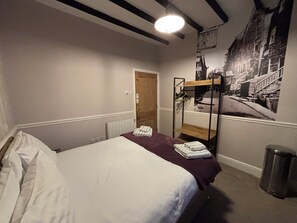 Een slaapkamer met een stapelbed, een radiator, een deur, een wandplank met kleding en een prullenbak.