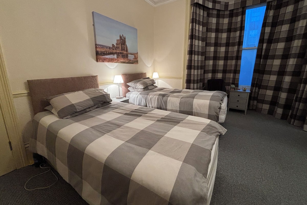A hotel room with two beds, a painting of a building, a window with curtains, and a desk with a chair.