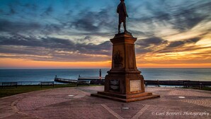 A statue on a pedestal with a plaque, set on a paved area with a view of the sea and a lighthouse in the background. - The Lansbury - Whitby  (Whitby)