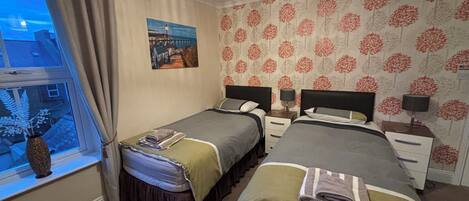 A bedroom with two beds, a window with curtains, a painting on the wall, and a patterned wallpaper.