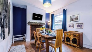 A dining area with a wooden table set for four, a brick fireplace, and a large window with curtains.