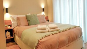 A neatly made bed with a green and pink bedspread, two bedside lamps, and a nightstand with a small plant. - King Studio 3 (Leiria)