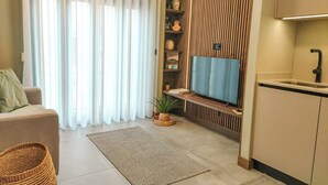 A modern living room with a sofa, a TV, a kitchen area with a sink, and a large window with sheer curtains. - King Studio 3 (Leiria)