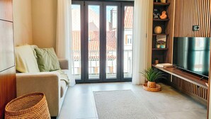 A modern living room with a sofa, a flat-screen TV, a large window with a view of buildings, and a wooden wall with shelves. - King Studio 3 (Leiria)