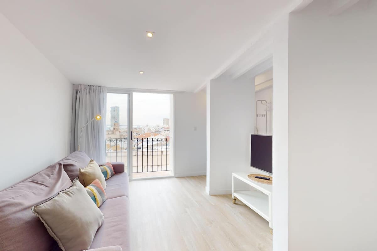 Een moderne woonkamer met een bank, een televisie op een standaard, en een balkon met uitzicht over de stad.
