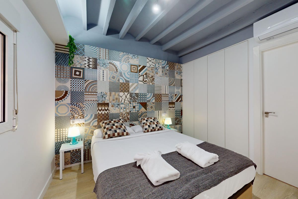 Een slaapkamer met een behang, een bed met kussens, een nachtkastje met een lamp en een deur.