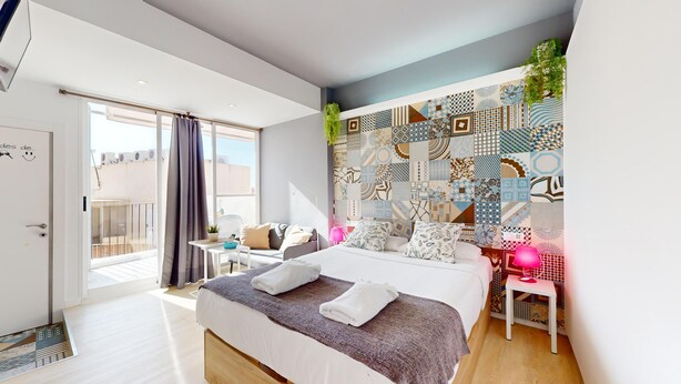 A modern bedroom with a large bed, a small desk, a balcony, and a patterned wall.
