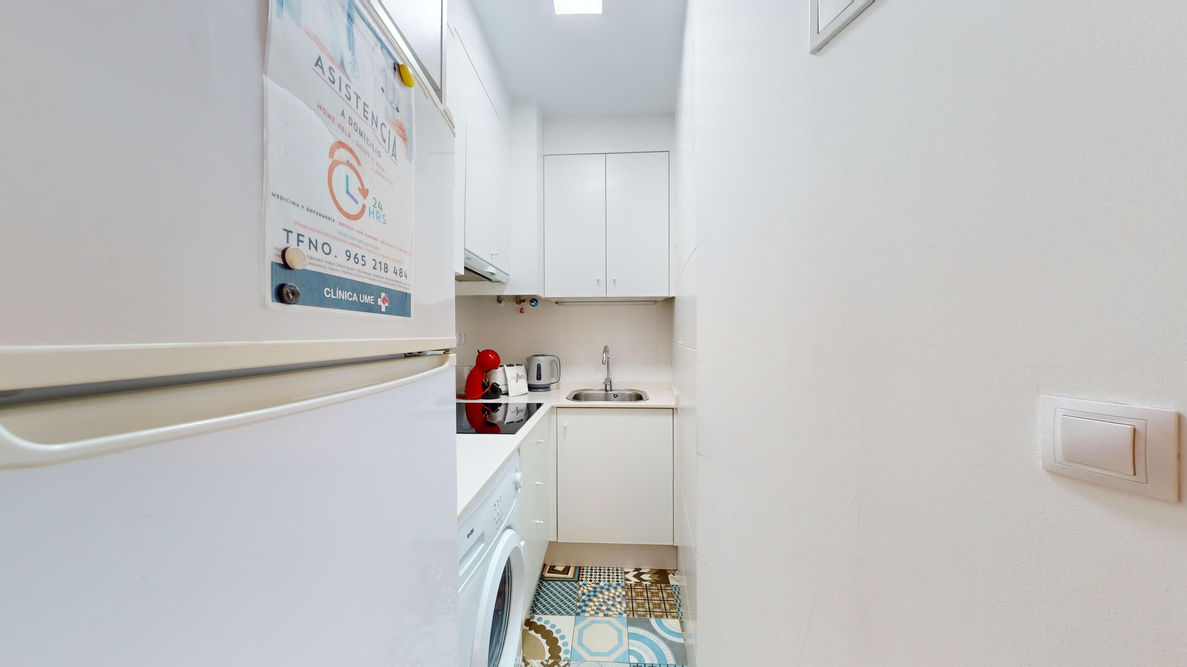 A narrow kitchen with white cabinets, a stainless steel sink, and a tiled backsplash. A refrigerator is on the left, and a washing machine is visible in the corner.