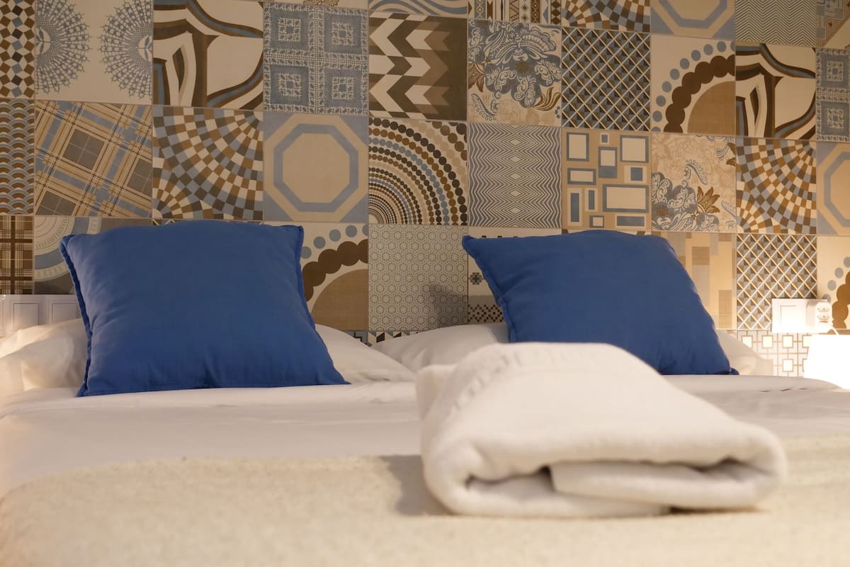 Een net opgemaakt bed met blauwe kussens en een opgevouwen witte handdoek, voor een muur met verschillende behangtegels.