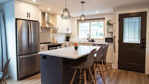A modern kitchen with stainless steel appliances, a central island with bar stools, pendant lighting, and a view of the outdoors through a window.