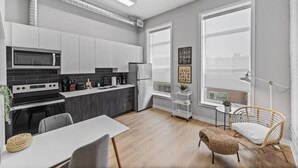 Une cuisine moderne avec des meubles blancs, une cuisinière noire et un réfrigérateur en acier inoxydable. On y trouve une table à manger avec des chaises, ainsi qu’un fauteuil en osier près de la fenêtre.