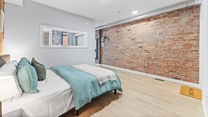 A bedroom with a bed, a bedside table, a mirror, a brick wall, and a wooden floor. - Stylish Loft Downtown w/ Views + Near River/Trails (Peterborough)