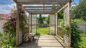 Puinen pergola, jossa on penkki ja koristeelliset säleikköpaneelit, ja jonka ympärillä on vehreää kasvillisuutta ja aita.