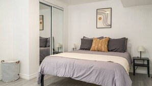 A bedroom with a bed, a bedside table, a lamp, a mirror, and a striped bag.