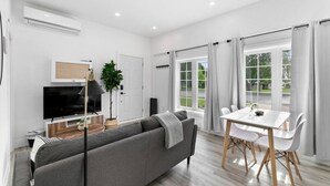 Una sala de estar moderna con un sofá gris, un televisor, una mesa de comedor con sillas y ventanas amplias con cortinas.
