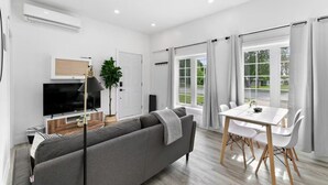 A modern living room with a grey sofa, a flat-screen TV, a dining table with white chairs, and large windows with curtains.