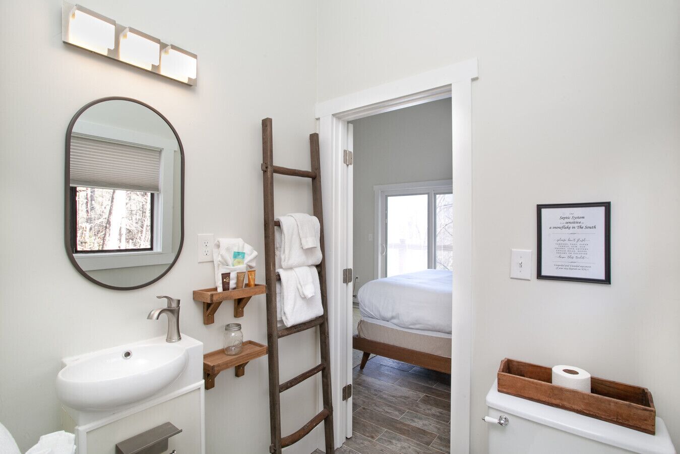 A bathroom with a sink, mirror, and wooden towel rack. A ladder leads to a bedroom with a bed and a window.