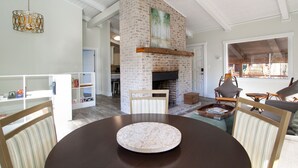 A spacious living room with a brick fireplace, a dining area with a round table, and a view of the outdoors through large windows. - Peaceful Heights with Cozy Decks and Smart TV! (Townsend)