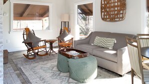 A spacious living area with a high ceiling, a grey sofa, wooden rocking chairs, a woven basket, and a patterned rug. - Peaceful Heights with Cozy Decks and Smart TV! (Townsend)