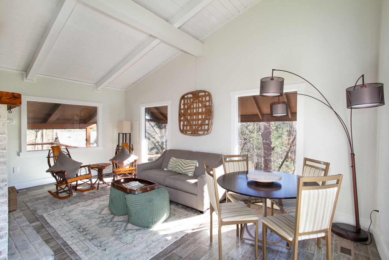 A cozy living room with a sofa, chairs, a coffee table, and a dining table. There are windows with views of trees, a floor lamp, and a wall-mounted light fixture.