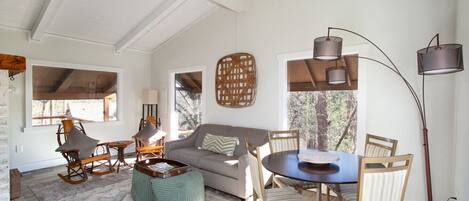 A cozy living room with a sofa, chairs, a coffee table, and a dining table. There are windows with views of trees, a floor lamp, and a wall-mounted light fixture.