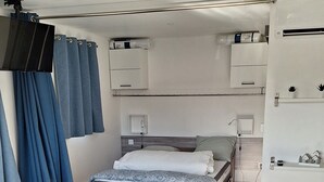 Quarto compacto e bem iluminado, com cama, mesas de cabeceira, escrivaninha e armário.