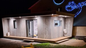 Um edifício moderno com um letreiro de neon que diz "Drive" e uma pequena área externa com mesas e cadeiras amarelas.