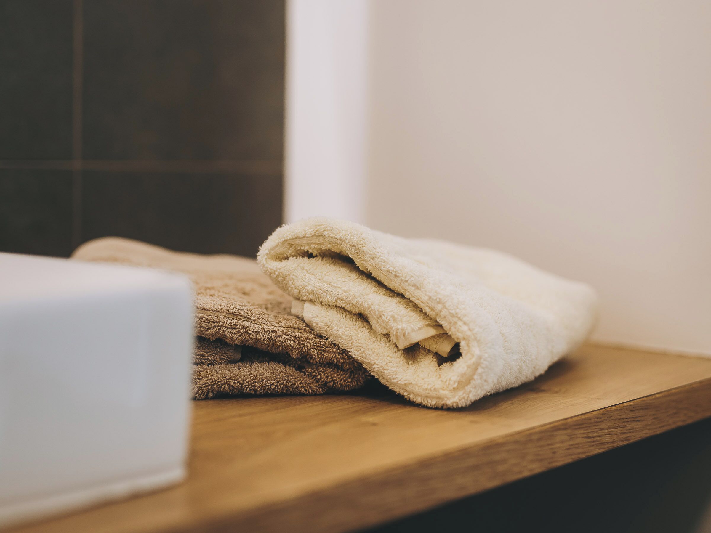 A wooden shelf with folded towels and a white object, likely a soap dispenser, in a bathroom setting.