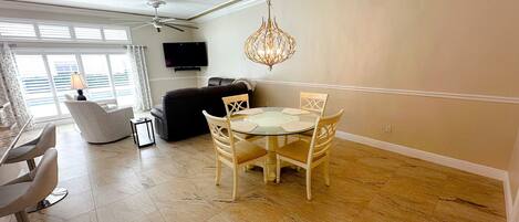 A modern living room with a dining area, a flat-screen TV mounted on the wall, and a chandelier.