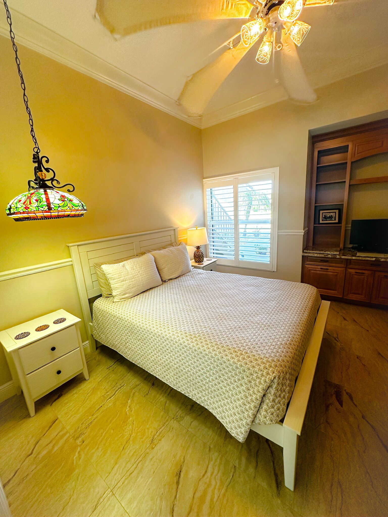 A bedroom with a bed, nightstands, a lamp, a ceiling fan, and a window with blinds.
