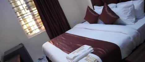 A hotel room with a bed, a window with blinds, a nightstand, and a wall-mounted heater.