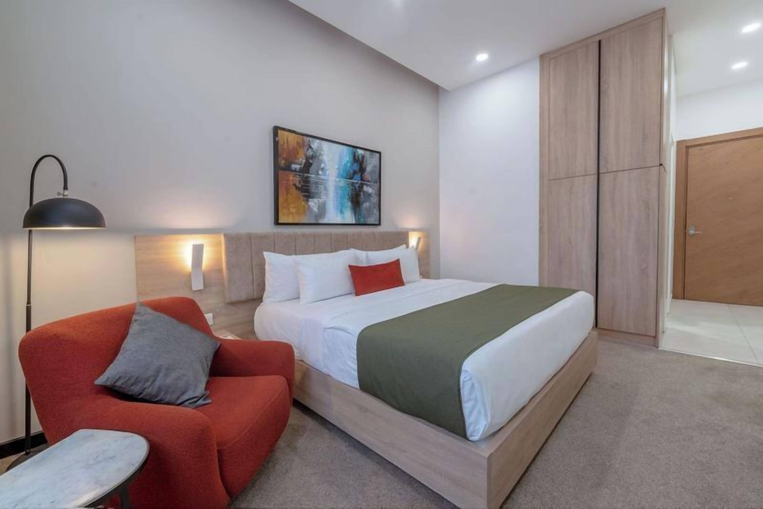 Una habitación de hotel moderna con una cama, un sillón rojo, una mesita, una lámpara de pie y un cuadro en la pared.