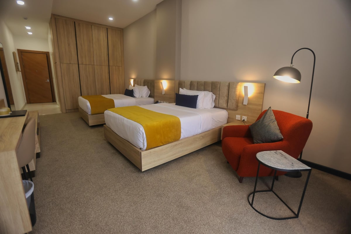 A hotel room with two beds, a desk, a chair, and a lamp.