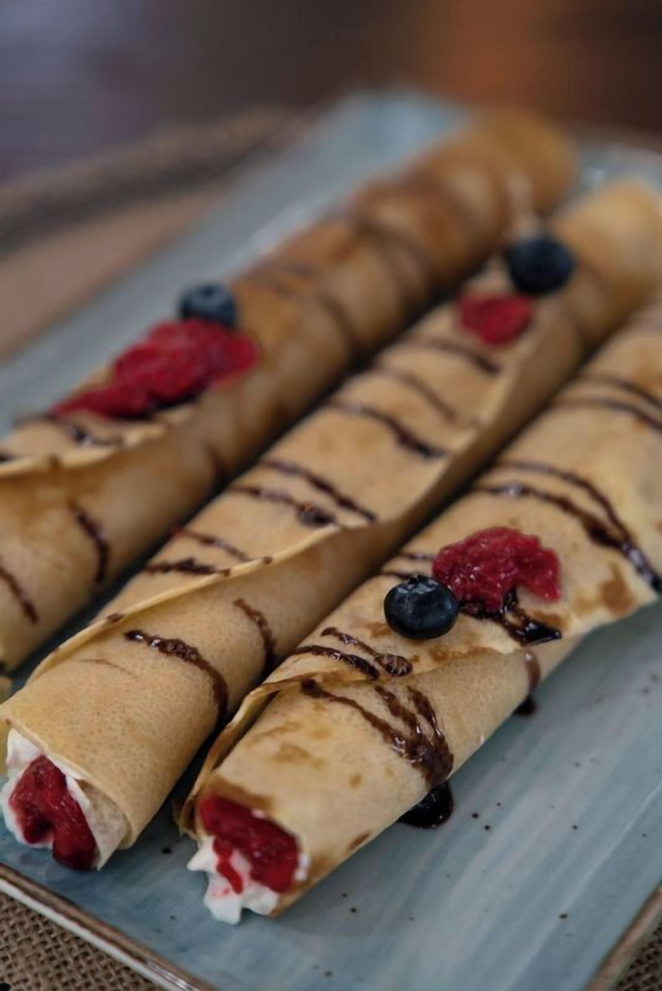 Tre crepes arrotolate con glassa al cioccolato e frutti di bosco su un piatto.