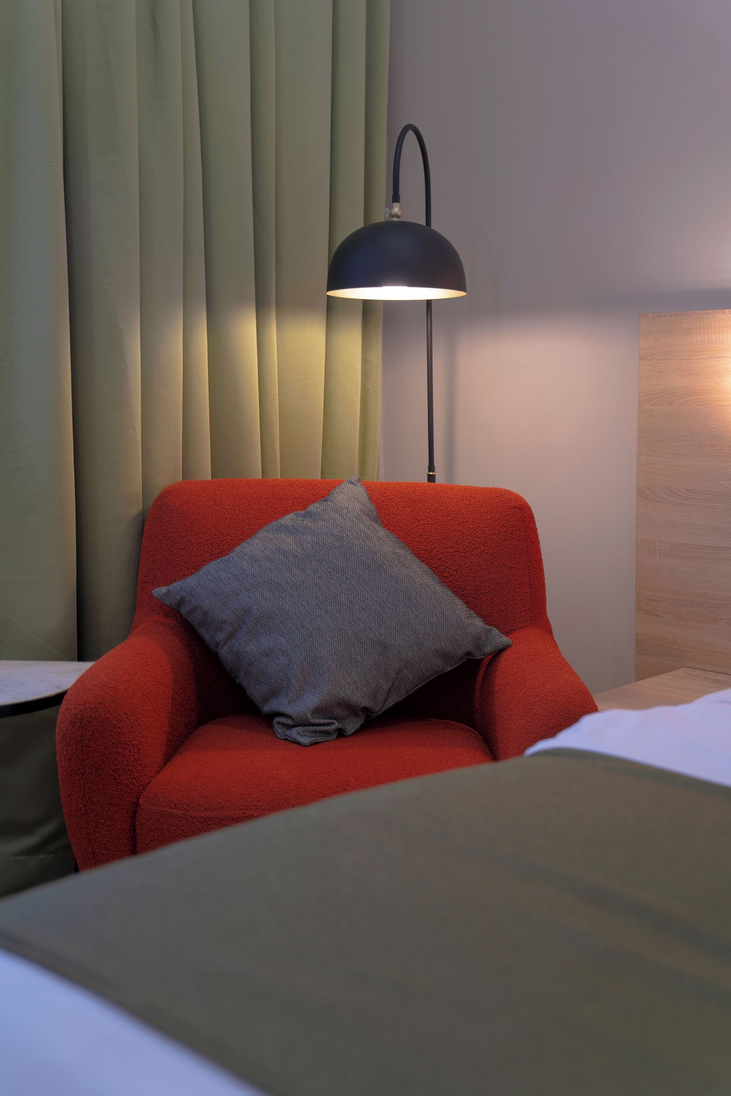 Een rode fauteuil met een grijze kussen, een zwarte vloerlamp en een houten hoofdbord.