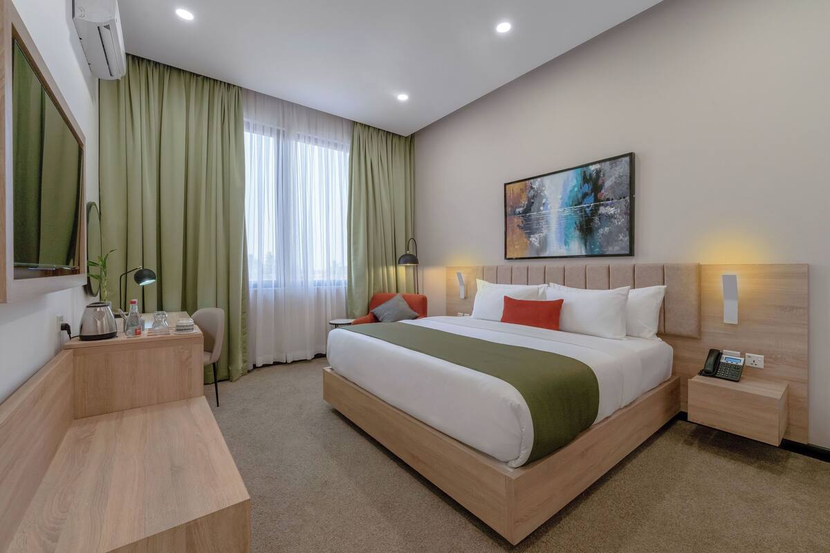 A hotel room with a large bed, a desk, a chair, and a bedside table with a phone.