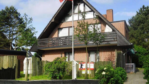 Een gebouw met twee verdiepingen, een schuine kap, een vlagmast met vlag, en een houten omheining.