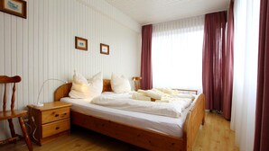 A wooden bed with white bedding, a bedside table, a chair, and a window with curtains. - Ferienwohnung in Westerland-ex TUI (Sylt)