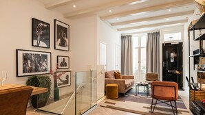 A modern living room with a glass partition, a sofa, a chair, and a coffee table. There are framed pictures on the wall and a bookshelf with decorative items.