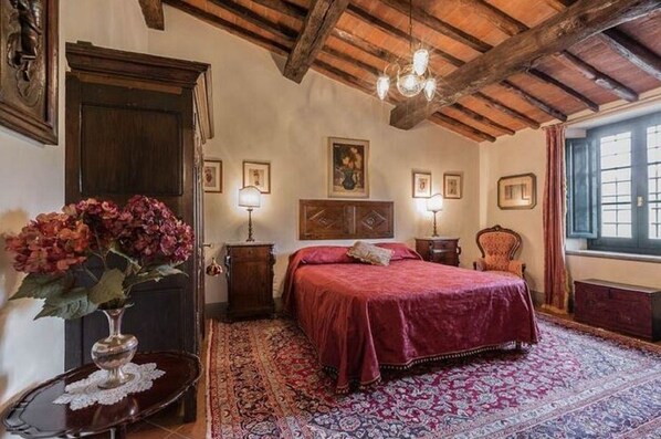 Un dormitorio con techo de madera, una cama grande con colcha roja, una mesita de noche de madera, una silla y una ventana con cortinas.