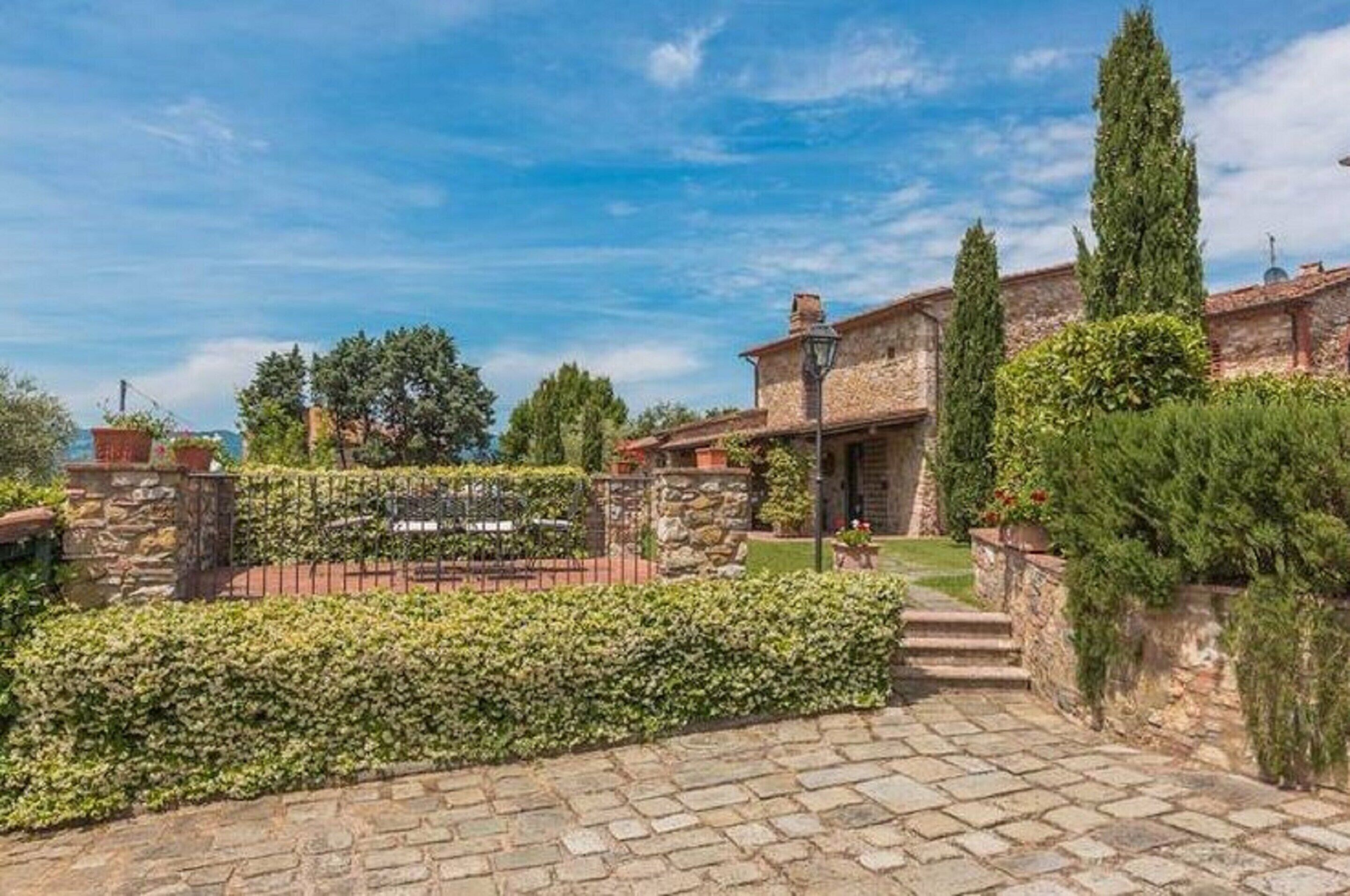 A stone building with a paved entrance, surrounded by well-maintained hedges and a tall cypress tree.