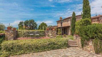 A stone building with a paved entrance, surrounded by well-maintained hedges and a tall cypress tree.