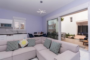 A modern living room with a sofa, patterned cushions, a dining area, and a sliding glass door leading to an outdoor patio.