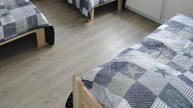 Habitación con tres camas individuales, cada una con una colcha de estampado diferente. Se observan mesitas de noche de madera con lámparas, un radiador y una ventana que proporciona luz natural.