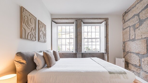 Un dormitorio con cama, dos ventanas, una pared de piedra y obras de arte en la pared.
