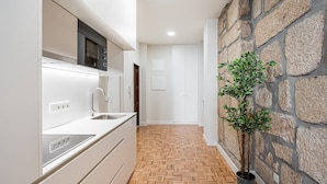 Una cocina moderna con armarios blancos, un microondas integrado y un lavamanos. El suelo tiene un diseño tipo espiga, y hay una planta en maceta junto a una pared de piedra.