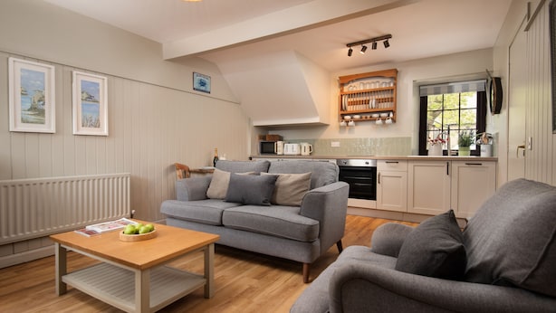 Ein modernes Wohnzimmer mit einem grauen Sofa, einem Holztisch und einer Küchenzeile mit weißen Schränken und einem Fenster.