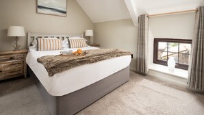 A bedroom with a large bed, a nightstand, a lamp, a window with curtains, and a painting on the wall. - One Bedroom Cottage in the heart of Beadnell (Beadnell)