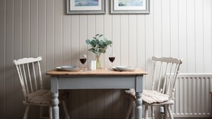 A dining table with two chairs, a vase with greenery, and two framed pictures on the wall. - One Bedroom Cottage in the heart of Beadnell (Beadnell)